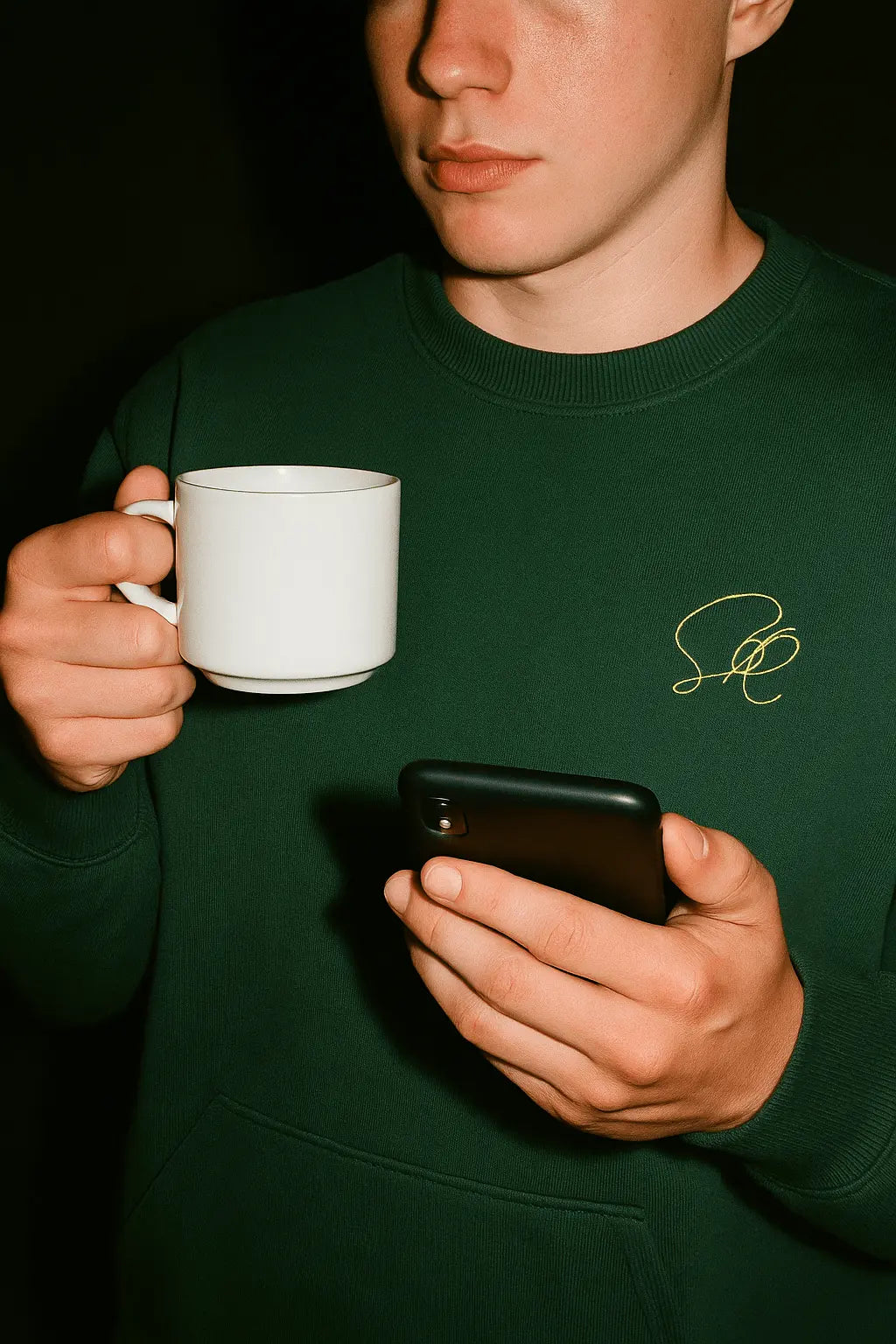 Person wearing a green sweatshirt with a logo, holding a white mug and a phone.