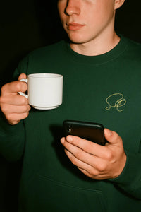 Person wearing a green sweatshirt with a logo, holding a white mug and a phone.