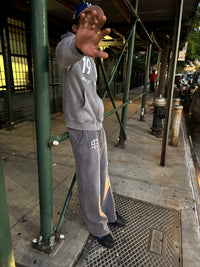 Person in grey90 streetwear hoodie and pants standing on a sidewalk with a green metal structure.